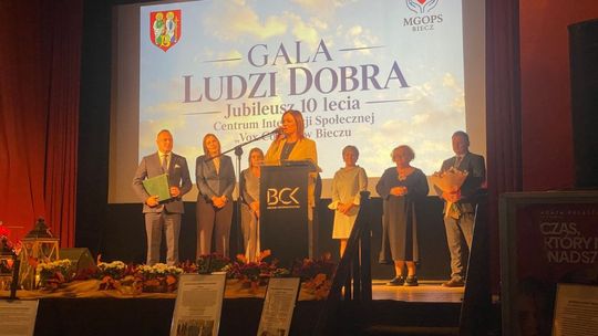 kobieta na scenie przed mikrofonem a za nią grupka ludzi