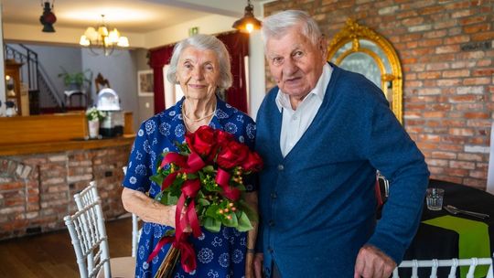 zdjęcie przedstawia portret starszej uśmiechniętej kobiety i mężczyzny, kobieta trzyma czerwone róże