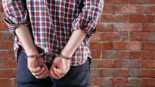 Afera edukacyjna w Małopolsce. Śledczy badają tropy z Nowego Sącza. Gorlice też zamieszane? zdjęcie przedstawia kajdanki nałożone na nadgarstki osoby, która stoi tyłem