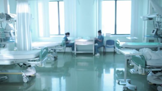 łóżka szpitalne po dwóch stronach sali w tle siedzące pielęgniarki przy stoliku rozmyte zdjęcie