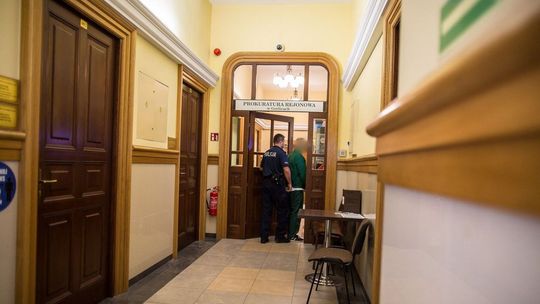 zdjęcie przedstawia policjanta, który prowadzi mężczyznę w zielonym stroju