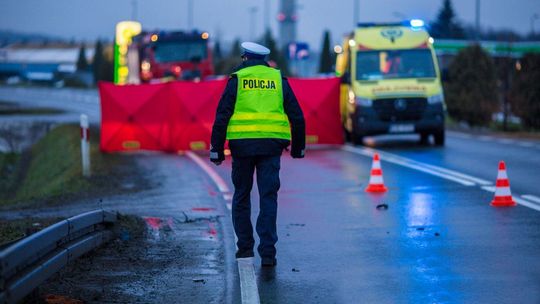 policjant widziany od tył, w tle czerwony parawan, karetka pogotowia i wóz straży pożarnej