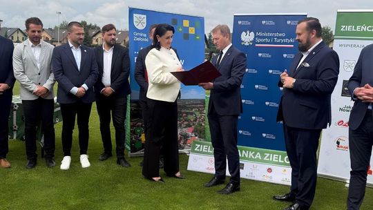 zdjęcie przedstawia grupę osób na boisku sportowym, za nimi banery reklamowe, mężczyzna trzyma przed sobą dokumenty w otwartej teczce