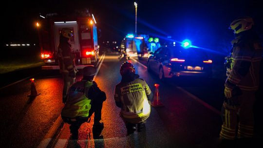 zdjęcie przedstawia strażaka i policjanta kucającego na jezdni, w tle samochody policyjne i straży pożarnej