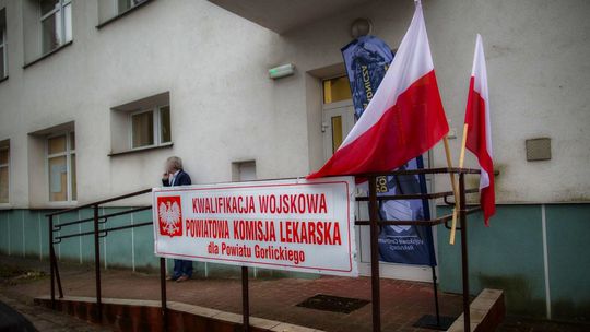 zdjęcie przedstawia wejście do budynku przed którymi jest baner informacyjny i flagi Polski