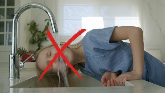 Dziecko pijące wodę z kranu, na pierwszym planie czerwony krzyżyk informujący o braku wody