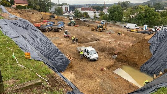 plac budowy na którym stoją auta, wywrotki, koparki i pracownicy, widok z daleka