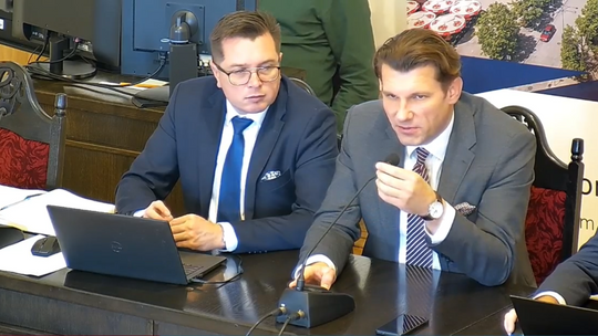 zdjęcie przedstawia dwóch mężczyzn pod krawatem i w garniturach siedzących przy stole
