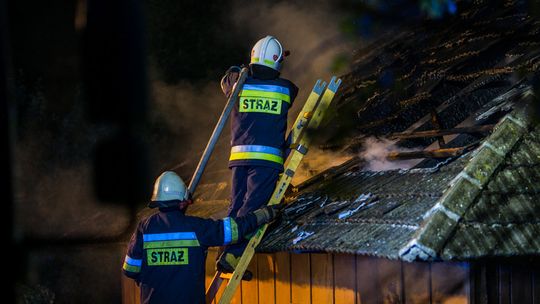 strażacy stojący na drabinie opartej o dach palącego się domu