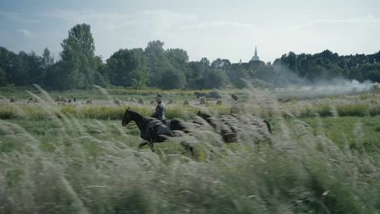 dwóch mężczyzn na koniach jadący wśród traw, w tle kościół wśród drzew