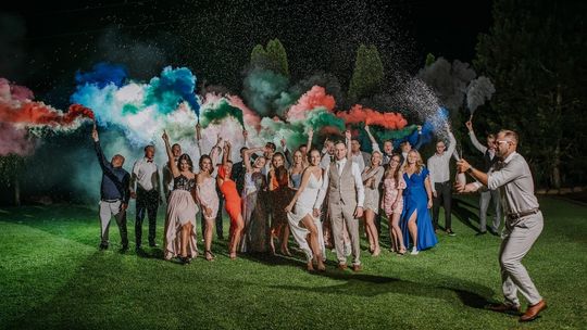 grupa ludzi bawiąca się kolorowymi proszkami podczas przyjęcia weselnego w ogrodzie w świetle ogni sztucznych
