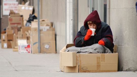 bezdomna osoba siedząca pod kocem na ulicy w otoczeniu kartonów