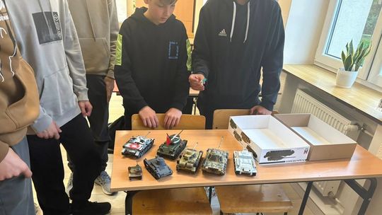 zdjęcie przedstawia małe modele czołgów stojących na ławce szkolnej, obok stoją chłopcy
