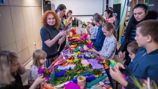 zdjęcie przedstawia kobietę i stół przy którym są dzieci i dorośli podczas warsztatów robienia palm wielkanocnych