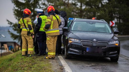 zdjęcie przedstawia czarny samochód stojący na drodze, obok policjant, strażacy i kilka osób