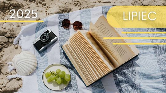Zdjęcie przedstawia koc na plaży z aparatem, kapeluszem i winogronami