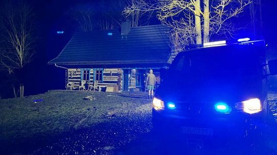 stojący radiowóz z niebieskim światłem, w tle drewniany dom, widok w nocy