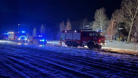 zdjęcie przedstawia wozy strażackie na drodze, w tle drzewa i dom drewniany