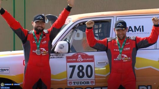 zdjęcie przedstawia dwóch mężczyzn w kombinezonach rajdowych na tle samochodu