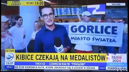 mężczyzna z mikrofonem, za nim stoi inny z flagą i napisem Gorlice - miasto światła