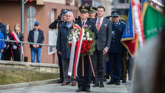 zdjęcie przedstawia grupę osób odświętnie ubranych, pierwsza z nich w mundurze niesie wieniec