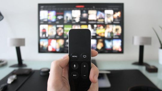 Zdjęcie przedstawia pilot telewizyjny w dłoni na tle telewizora