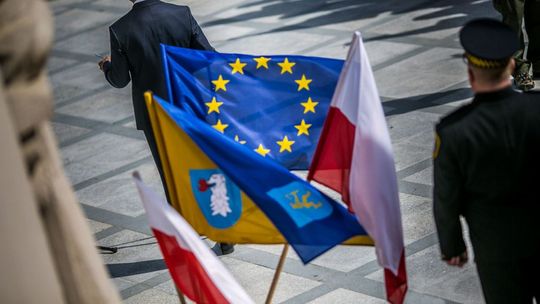 flagi unii europejskiej, gorlic i polski na stelażu, obok mężczyzna w mundurze strażnika miejskiego