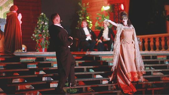 zdjęcie przedstawia scenę i kobietę w sukni wskazującą palcem na mężczyznę