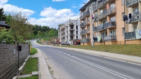 Zdjęcie przedstawia bloki mieszkalne przy ulicy, w oddali las