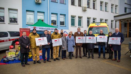 zdjęcie przedstawia grupę osób trzymających tabliczki, za nimi budynek i karetka