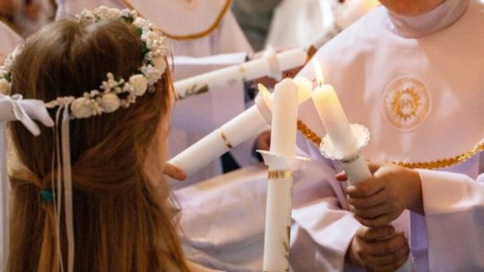 zdjęcie przedstawia dzieci pierwszokomunijne w białych strojach trzymające świece w rękach, odpalające sobie ogień