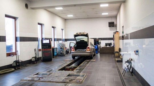 zdjęcie przedstawia wnętrze garażu z samochodem na środku i mechanikiem oraz urządzeniami