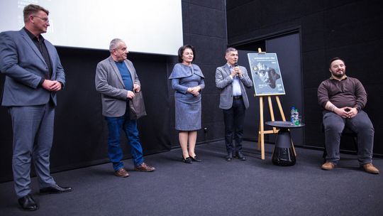 zdjęcie przedstawia osoby stojące przed ekranem w kinie, jedna osoba siedzi na krzesełku