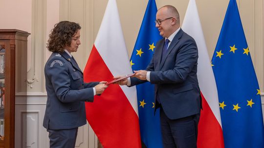 zdjęcie przedstawia kobietę w mundurze, której mężczyzna wręcza teczkę, w tle flagi państwowe