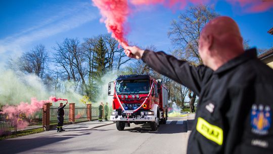 zdjęcie przedstawia mężczyznę trzymającego racę, obok przejeżdża samochód strażacki