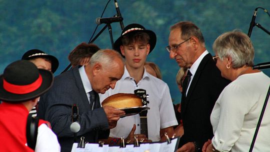 mężczyzna w garniturze pochylający się nad chlebem, żeby go ucałować podczas dożynek, obok inne osoby przyglądają się temu