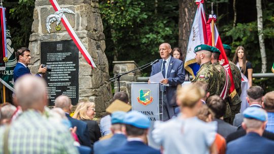 mężczyzna mówiący do mikrofonu przy mównicy przed grupą osób, obok pomnik w krzywej banicy
