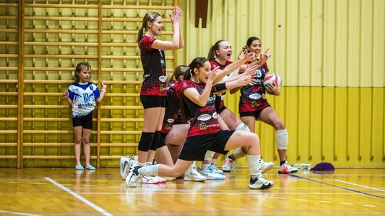 zdjęcie przedstawia dziewczyny w czerwonych strojach sportowych pokazujących palcami w stronę boiska
