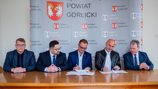 zdjęcie przedstawia mężczyzn w garniturach siedzących przy stole podpisujących dokument