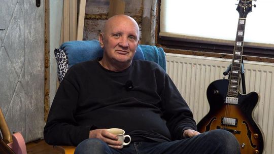 zdjęcie przedstawia mężczyznę z filiżanką w ręce, obok gitara