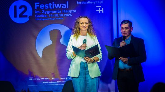 kobieta w jasnym garniturze i męczyzna stojący na scenie, obok baner z napisem 12 festiwal