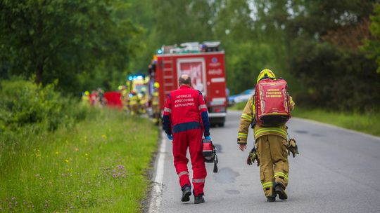 zdjęcie przedstawia dwóch mężczyzn w kombinezonach idących jezdnią, w tle wóz strażacki i karetka