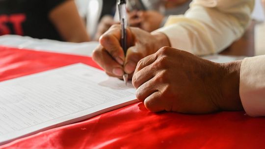 zdjęcie przedstawia rękę w której znajduje się długopis podpisujący dokument na stole z czerwonym obrusem