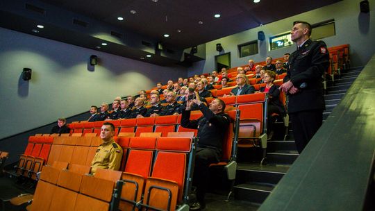 sala wypełniona osobami siedzącymi z fotelach obok stoi mężczyzna w mundurze galowym straży pożarnej