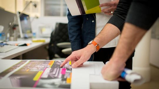 zdjęcie przedstawia dłonie grafików położone na biurku wskazujące na materiały graficzne
