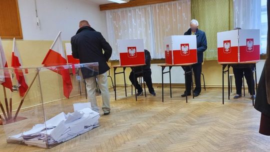 Sala głosowania w gorlickim lokalu wyborczym, mężczyzna za parawanem wyborczym, obok urna i flagi państwowe