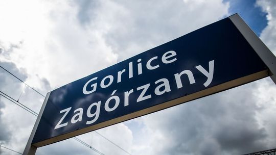 Tablica na stacji PKP Gorlice-Zagórzany w tle zachmurzone niebo i przewody trakcji kolejowej