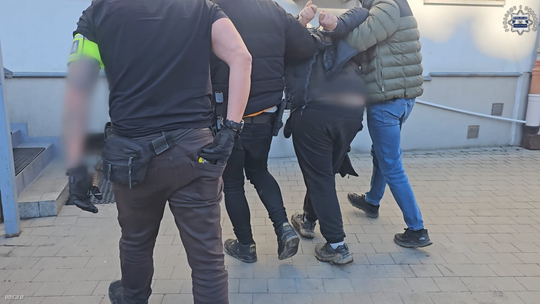 zdjęcie przedstawia policjantów prowadzących zatrzymanego w kajdankach