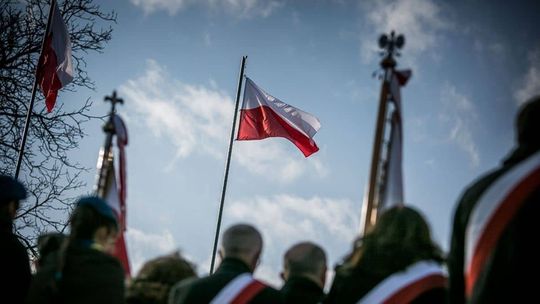 na pierwszym tle sylwetki osób, w centralnym punkcie obrazu polska flaga na maszcie na tle błękitnego nieba