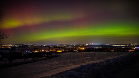 zdjęcie przedstawia kolorowe smugo na niebie, w tle rozświetlone światła miasta